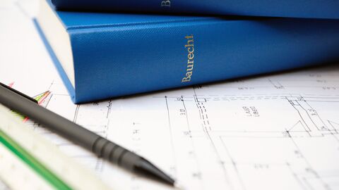 Auf einem Tisch liegen zwei blau eingebundene Bücher übereinander, auf dem Einband steht in goldener Schrift Baurecht. Die Bücher liegen zusammen mit einem braunen Kugelschreiber auf einer schwarz-weißen Zeichnung.