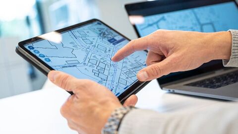 Person hält Tablet mit Bauplan Eine Person hält ein Tablet in der einen Hand und zeigt mit der anderen Hand darauf. Auf dem Bildschirm ist ein Bauplan in schwarz-weiß zu sehen.