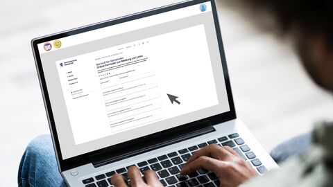 Eine Person hat ein aufgeklapptes Laptop auf den Beinen liegen und die Hände auf der Tastatur. Auf dem Bildschirm ist ein Online-Formular mit ausfüllbaren Feldern zu sehen.