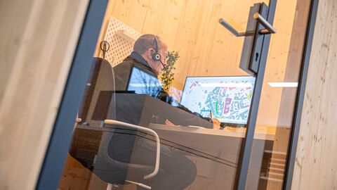 Arbeit an Bebauungsplan Ein Mann sitzt in an einem Schreibtisch in einem Raum mit Glastür. Er hat ein Headset auf dem Kopf und schaut auf einen Bildschirm, auf dem ein Bauplan zu sehen ist.