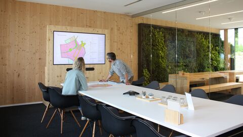 Teambesprechung vor Bildschirm In einem Besprechungsraum mit Holzvertäfelung und Glaswand sitzen zwei Personen auf schwarzen Stühlen an einem langen, weißen Konferenztisch und blicken auf einen Bildschirm an der Wand, Auf dem Bildschirm ist ein Bebauungsplan zu sehen. Rechts im Bild sind Gemeinschaftsräume mit Begrünung zu sehen.