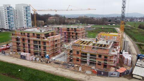 Rohbau von vier Mehrfamilienhäusern. Baumaterial und Bauzäune, 2 Baukräne. Ein Mann steht auf der Ladefläche eines LKW. Zwei Hochhäuser stehen links im Bild. Ringsherum ist Wiese und am oberen Bildrand eine Stadt, Wald und grauer Himmel.