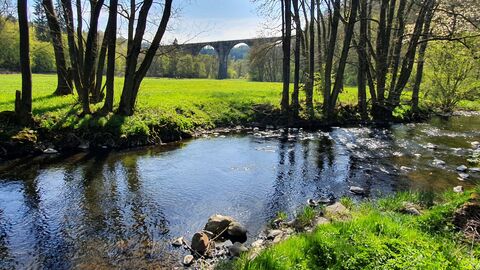 Bachlauf Ein breiter flacher Bachlauf. Am Ufer stehen Bäume und es wächst Gras. Im Hintergrund ist eine grüne Wiese und Teile eines Viaduktes mit Rundbögen zu sehen. Dahinter ist ein Wald unter blauem Himmel mit einzelnen Wolken