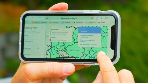 Interaktive Karte auf Smartphone Eine Person hält ein Smartphone in der einen Hand. Mit dem Zeigefinger der anderen Hand zeigt sie auf das Display eines Smartphones. Auf dem Display ist eine grün schattierte Landkarte zu sehen.